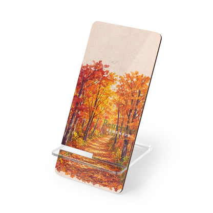 A rectangular printed PHONE STAND with rounded corners, shown standing on a clear acrylic display stand against a white background. The printed design depicts an autumn forest path with orange and red foliage under a pale sky, and the words 'Remember' are faintly visible near the center.