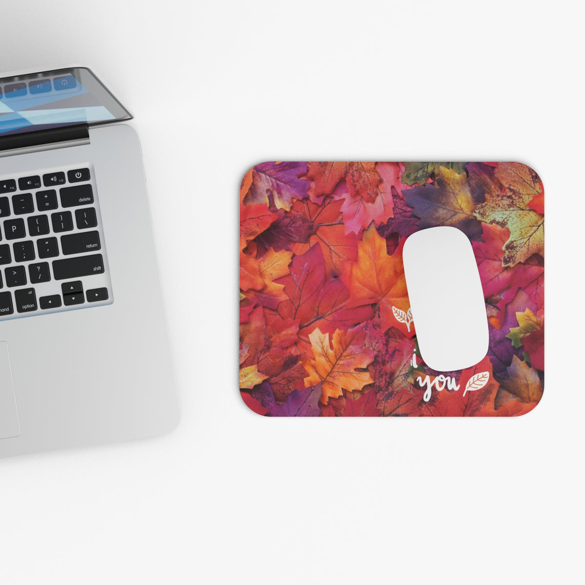 A rectangular computer mouse pad with a colorful autumn leaf pattern, shown on a white desk next to a silver laptop. The mouse pad features a white cutout for the mouse and the word 'you' printed in white script with small leaf details.