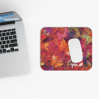 A rectangular computer mouse pad with a colorful autumn leaf pattern, shown on a white desk next to a silver laptop. The mouse pad features a white cutout for the mouse and the word 'you' printed in white script with small leaf details.