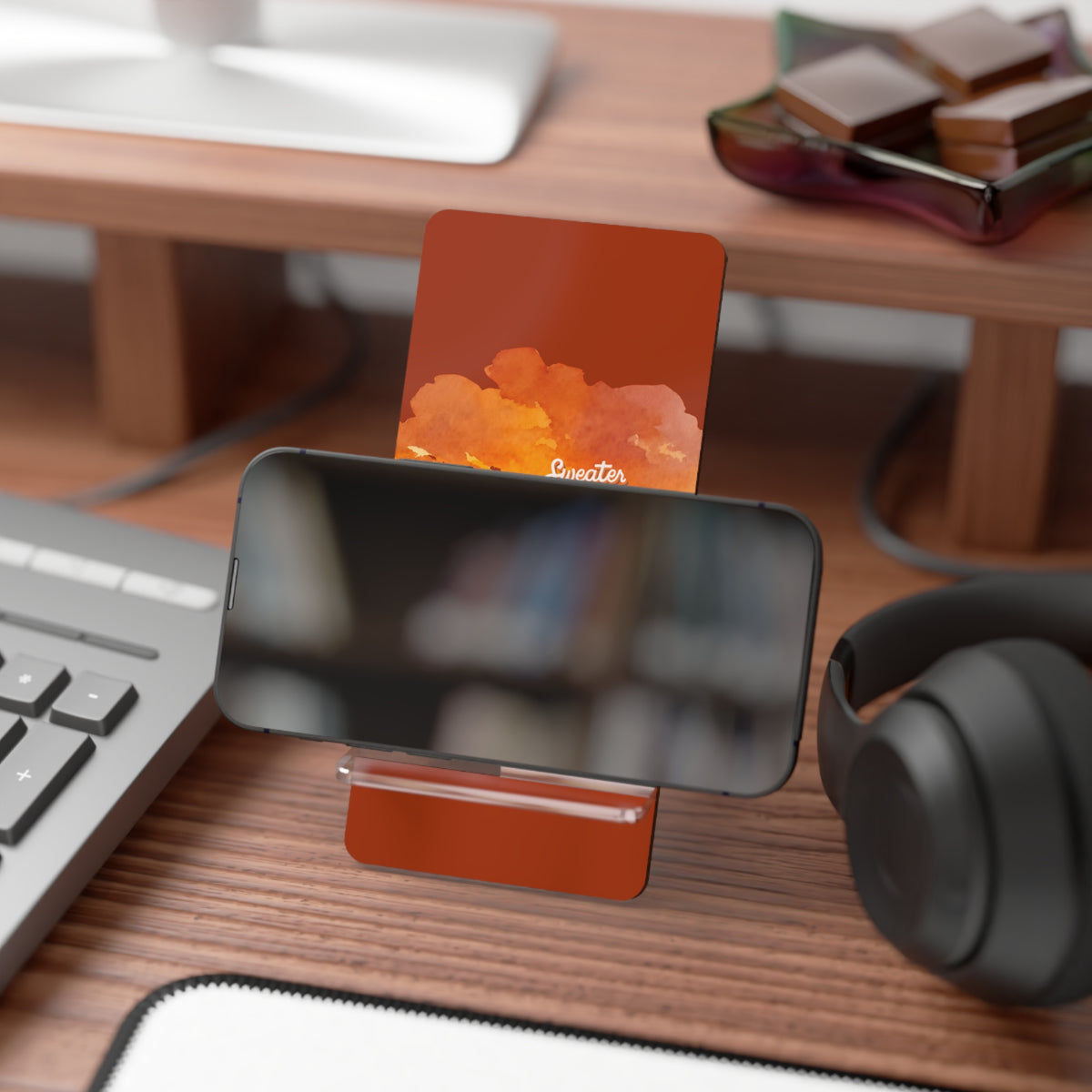 A modern PHONE STAND holding a smartphone and a card, placed on a wooden desk with a keyboard, headphones, and other office items in the background.