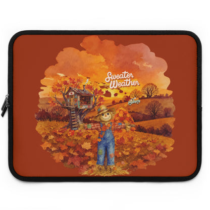A laptop sleeve featuring an autumn-themed illustration with a scarecrow, treehouse, fall leaves, and the words 'Sweater Weather' on a rust-orange background.