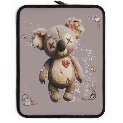 A laptop sleeve featuring a graphic print of a creepy, stitched teddy bear with a red heart on its chest, set against a dusty purple background with small skull and floral doodles. The sleeve has a black border and a black zipper on the left side.