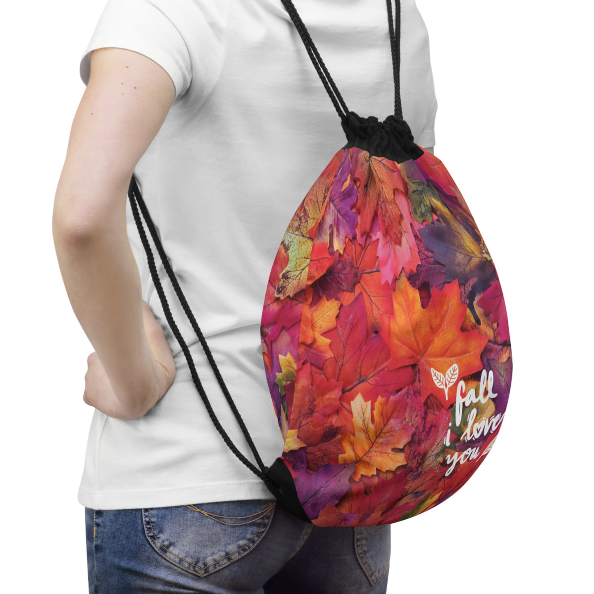 A person wearing a white t-shirt and blue jeans carries a drawstring backpack with a vibrant autumn leaf print. The backpack features a colorful pattern of red, orange, yellow, and purple maple leaves, with black drawstrings and white text reading 'fall I love you'.