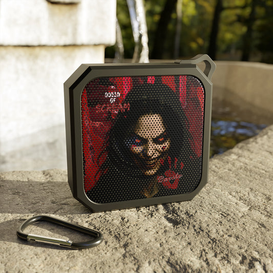 A rugged portable speaker with a dark gray frame and a red graphic featuring a creepy woman's face, placed on a stone surface outdoors with a metal carabiner nearby.
