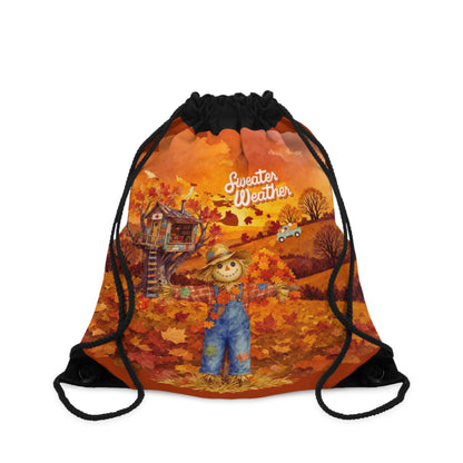 A drawstring backpack with a black top and black cords, featuring a colorful autumn landscape graphic with a scarecrow, treehouse, fall leaves, and the words 'Sweater Weather'.
