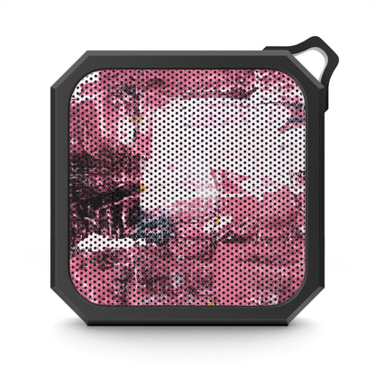 A rugged, square-shaped portable speaker with a black protective frame and a hanging loop, featuring a pink and white abstract patterned speaker grille on a white background.