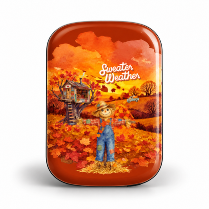 A glossy, MAGNETIC POWER BANK with a vibrant autumn-themed illustration. The design features a scarecrow in blue overalls standing in a field of orange and red maple leaves, with a treehouse, a small car, and the text 'Sweater Weather' written in white script.